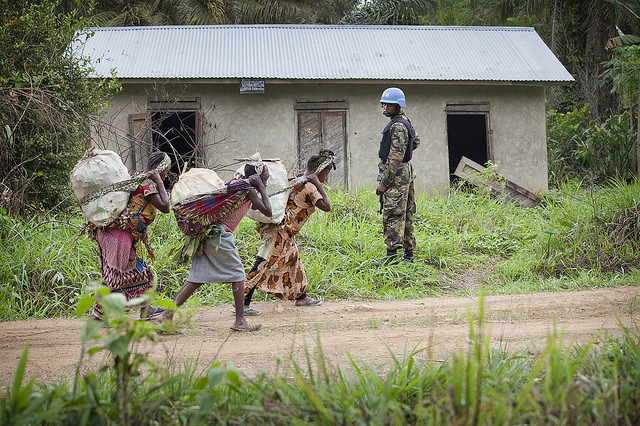 Women flee fighting in the Beni region, carrying their belongings past a soldier and an abandoned house 